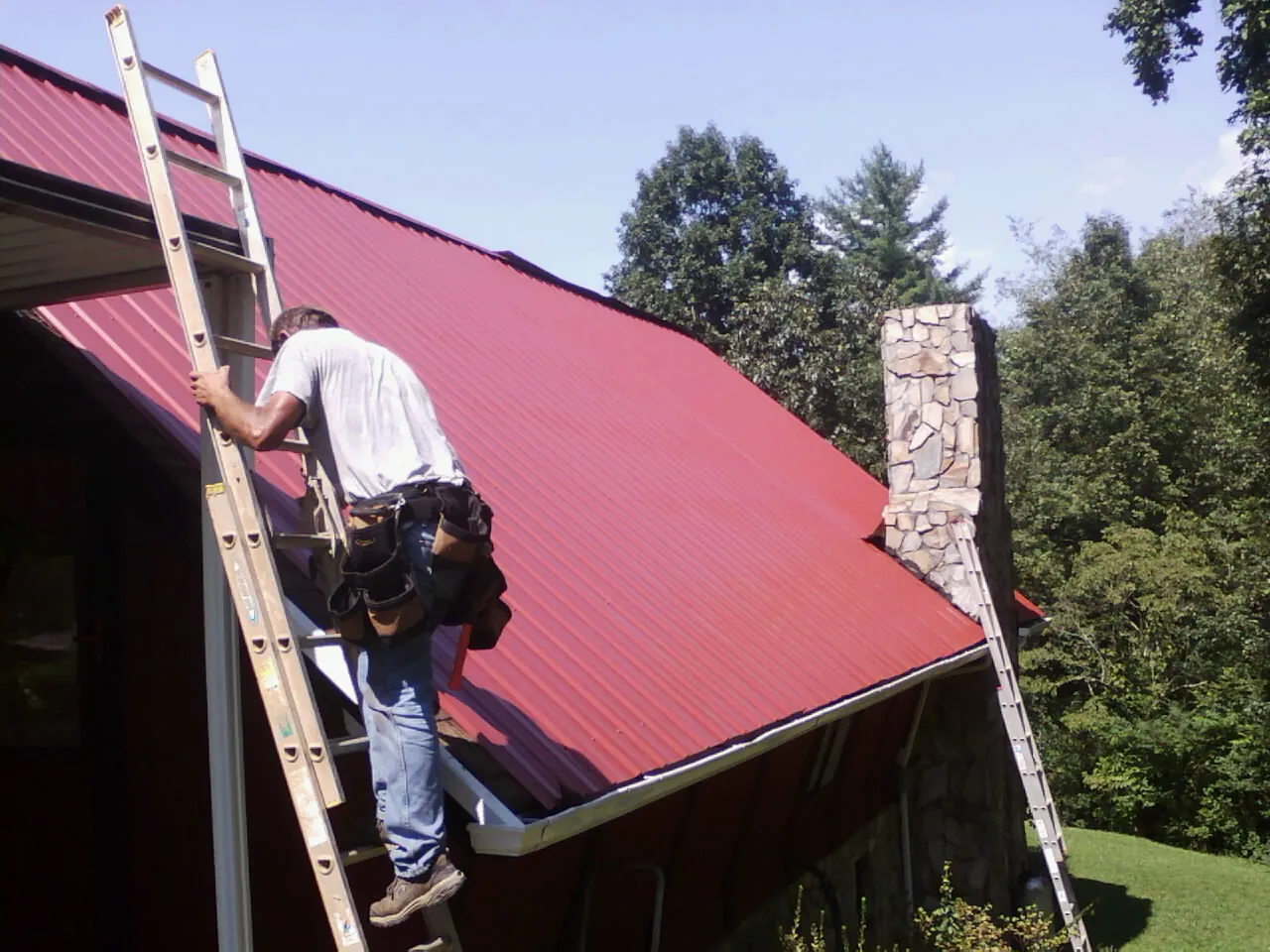 Roofer on a ladder beside a red metal roof and stone chimney — Soffit & Fascia Repair by Roofing Brothers in Bruin